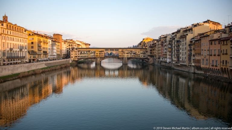 Long view down the Arno toward the shop-lined Ponte Vecchio, with riverside buildings reflected in calm water.