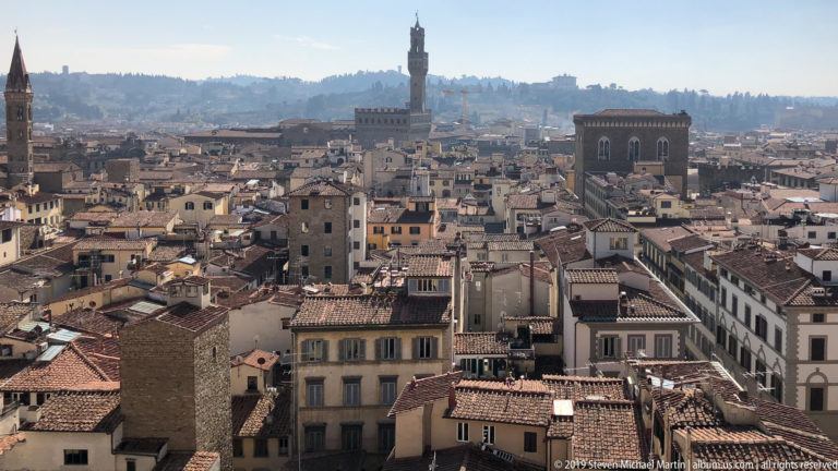 Telephoto view over Florence rooftops toward the Torre di Arnolfo of Palazzo Vecchio and hazy hills beyond.