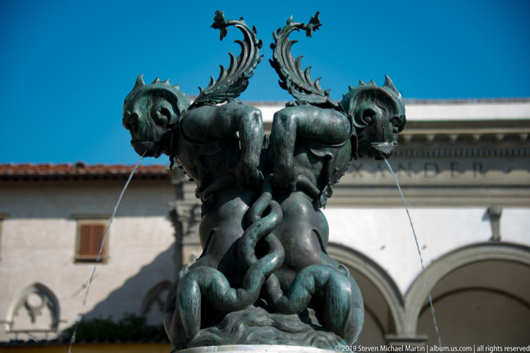 Bronze fountain with intertwined sea creatures, curling tails, open mouths, and thin streams of water.