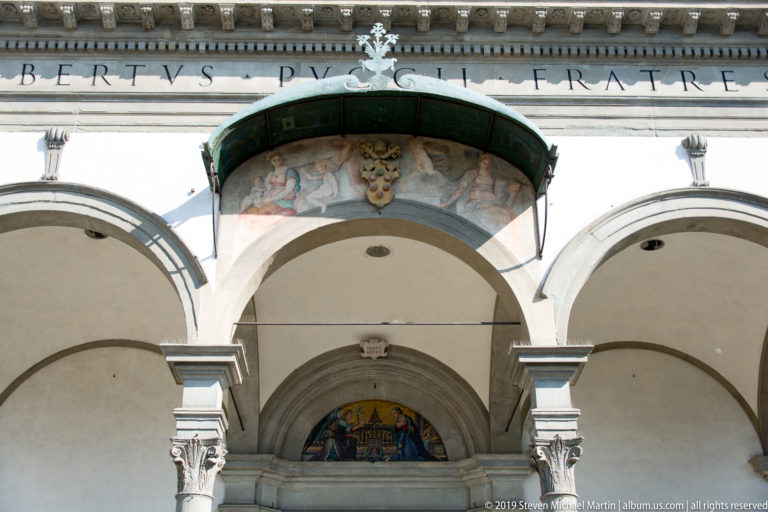 Central portico bay with broad arches, stone columns, inscribed entablature, painted lunette, and projecting canopy.