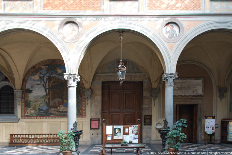 Three-arched cloister portico with stone columns, frescoes, portrait medallions, wooden doors, lantern, and notice boards.
