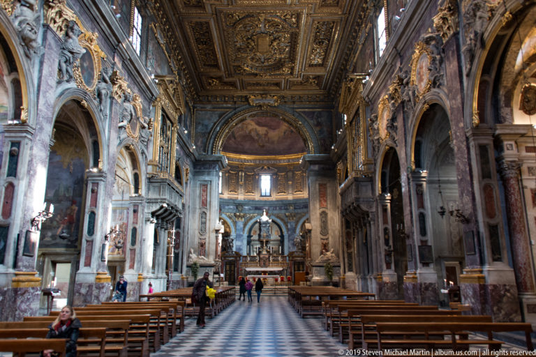 Wide symmetrical nave view toward sanctuary with pews, patterned aisle, marble arches, paintings, and gilded ceiling.