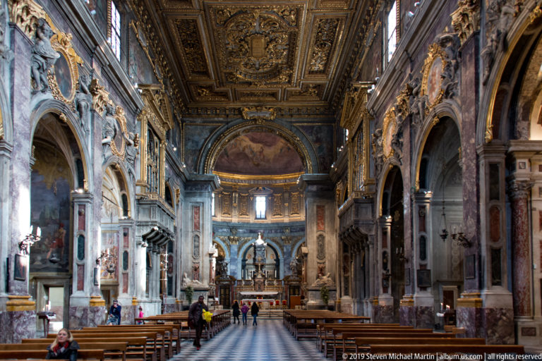 Centered nave with patterned aisle, pews, marble arcades, frescoes, sculptures, gilded ceiling, and distant altar.