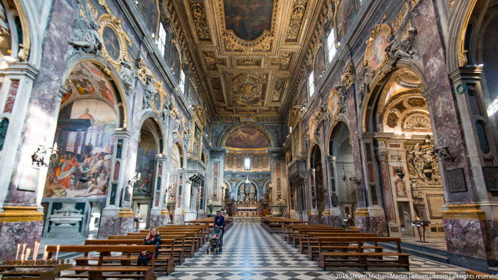 Central nave aisle with wooden pews, patterned floor, marble arches, frescoes, sculptures, and gilded ceiling.