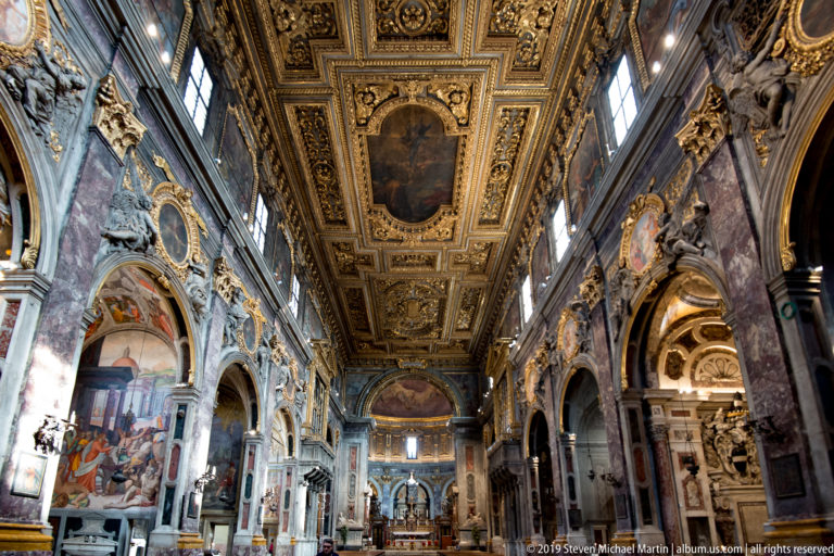 Oblique basilica nave with gilded ceiling, marble arches, frescoed walls, sculptures, and distant sanctuary.
