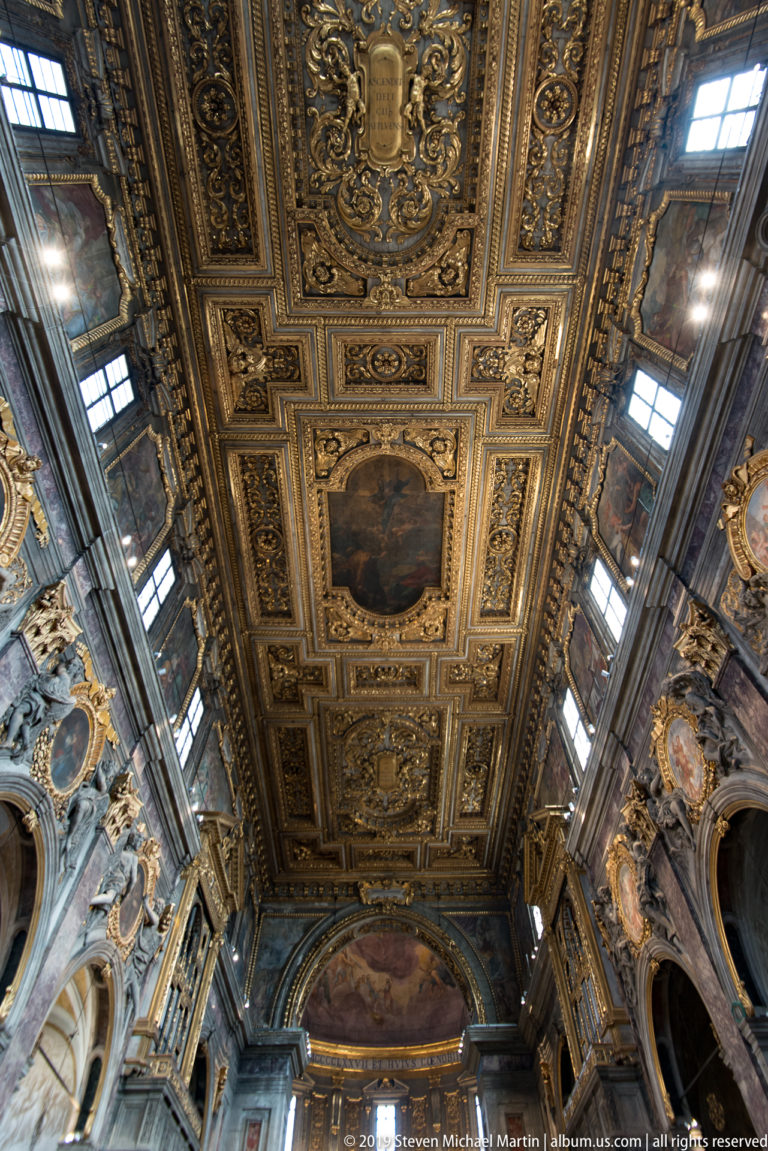 Steep upward view of gilded coffered nave ceiling with framed paintings, sculpted ornament, and side windows.