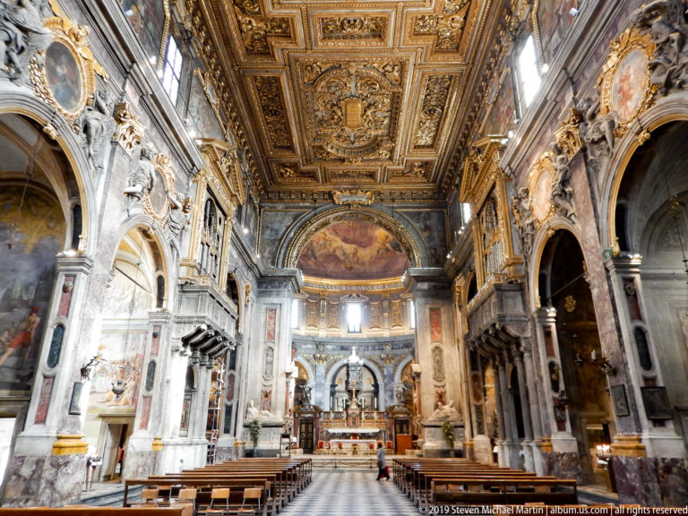 Formal axial nave view with patterned aisle, pews, marble arches, frescoes, sculptures, gilded ceiling, and sanctuary.