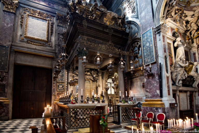 Ornate church altar beneath dark carved canopy with hanging lamps, candles, flowers, marble, and red chairs.