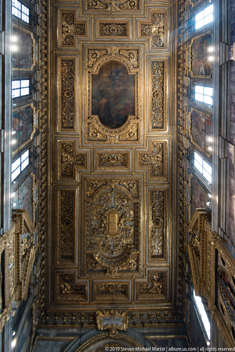 Near-vertical view of gilded coffered ceiling with framed paintings, sculpted ornament, and tall side windows.