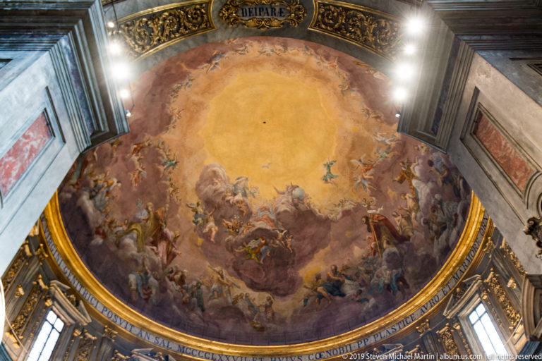 Steep upward view of frescoed dome with encircling inscription, drum windows, spotlights, and gilded border.