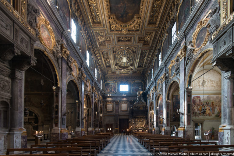 Centered church aisle with wooden pews, marble arches, frescoed walls, gilded ceiling, and bright distant window.