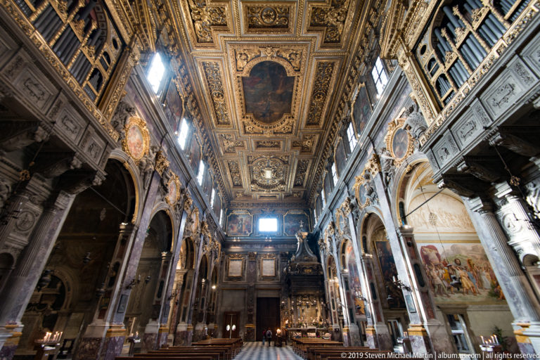 Long nave view beneath gilded coffered ceiling, with marble arcades, paintings, sculptures, frescoes, and high windows.