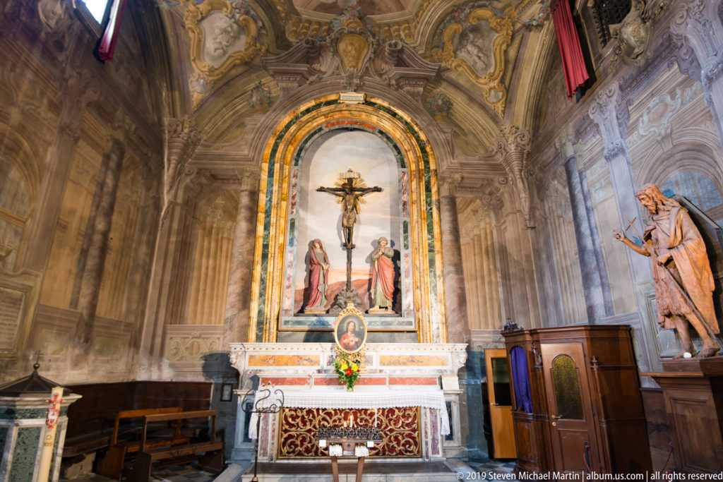 Side chapel with crucifix and flanking figures above a dressed altar, flowers, seating, and painted vault.