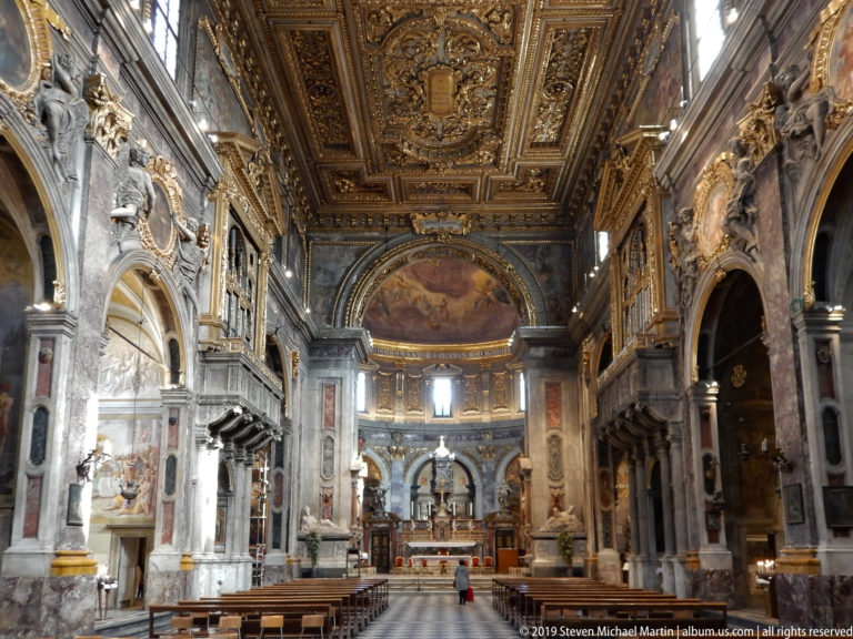 Wide basilica nave toward sanctuary with patterned aisle, pews, marble arcades, frescoes, sculptures, and gilded ceiling.