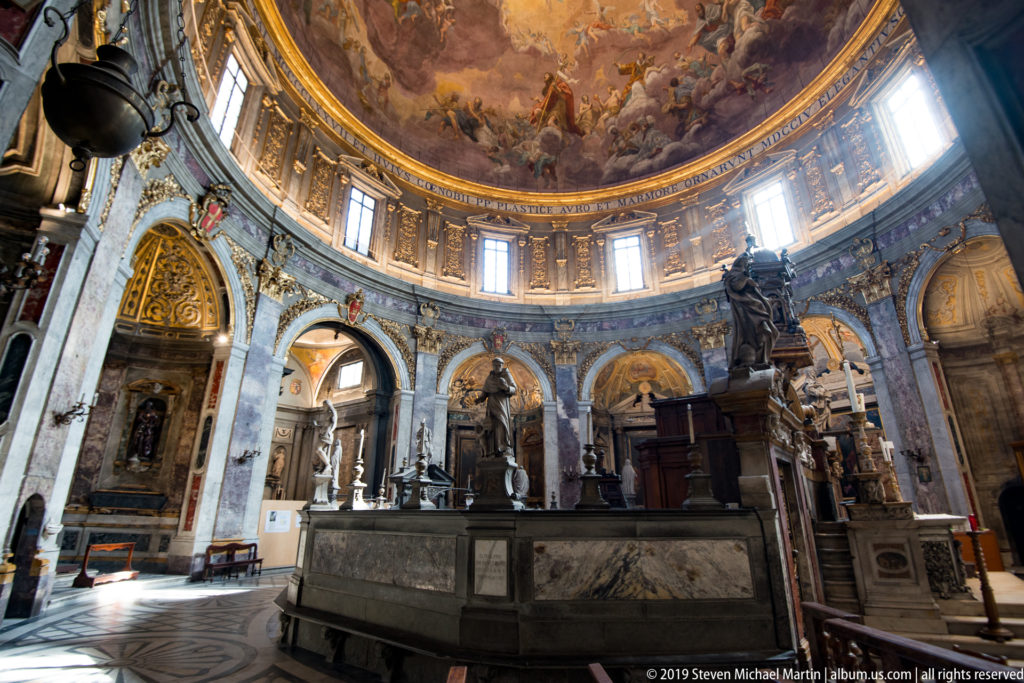 Wide domed church interior with marble arches, monumental sculptures, windows, and a large stone structure in the foreground.