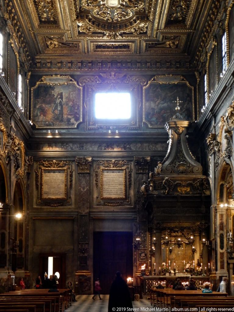 Dark church end wall with large paintings, gilded ceiling, bright window, doorway, and candlelit altar.
