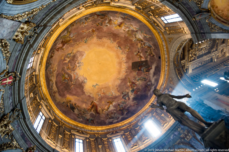 Vertical view of frescoed dome, bright drum windows, gilded details, and a dark statue at right.
