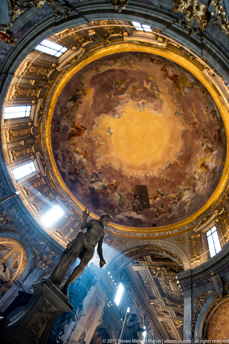 Oblique view of frescoed dome and bright drum windows above a dark foreground sculpture.