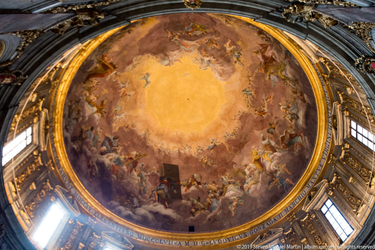 Centered frescoed dome with clouds and figures above an inscribed band, windows, and gilded architectural framing.