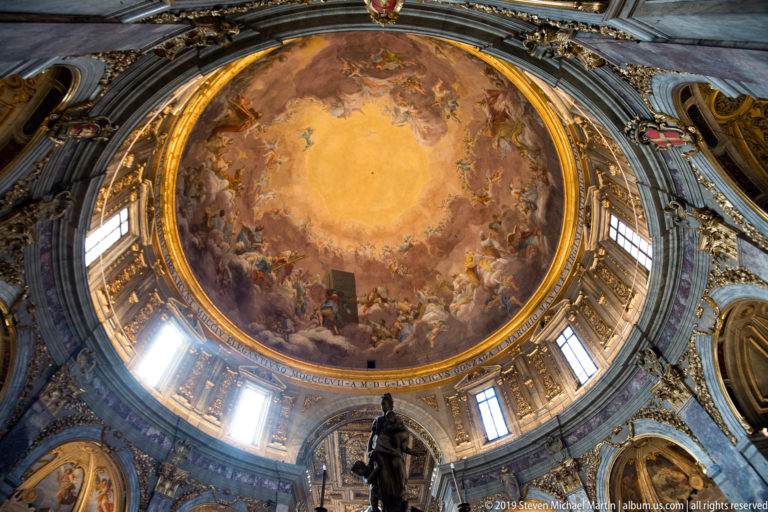 Upward view of a large frescoed dome with drum windows, gilded framing, and sculpture at the lower edge.