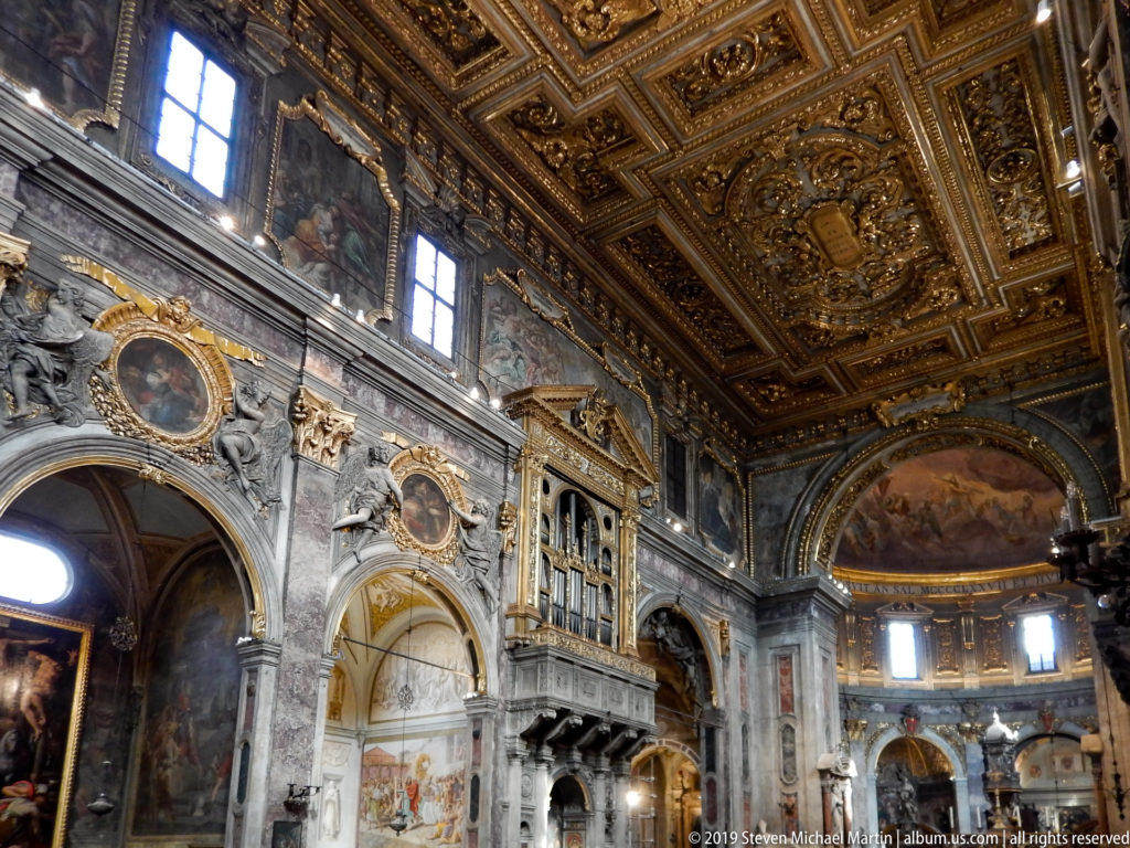 Wide basilica interior with marble arches, sculptures, frescoes, oval paintings, gallery, and gilded coffered ceiling.