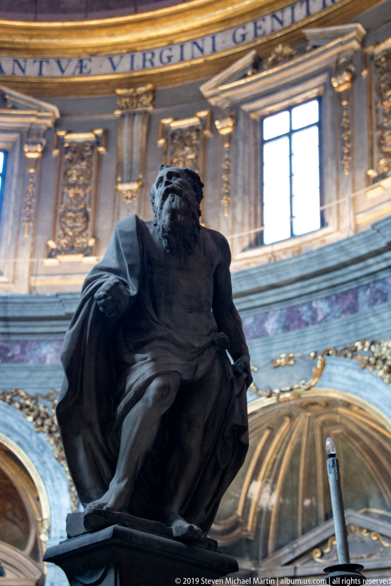 Dark seated male sculpture with heavy drapery against gilded panels, windows, and colored marble.