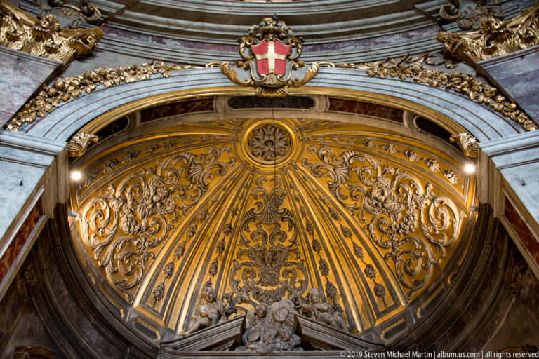 Gilded fan-shaped arch decoration with dark arabesques, radiating ribs, central rosette, and heraldic shield.