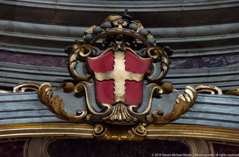 Red heraldic shield with a white cross, crowned and surrounded by sculpted scrollwork.