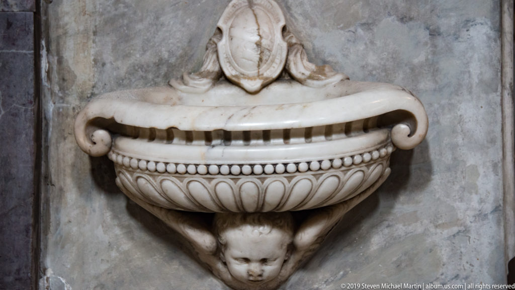 Wall-mounted marble basin with beaded molding, carved foliage, cherub head, and shield-shaped crest.
