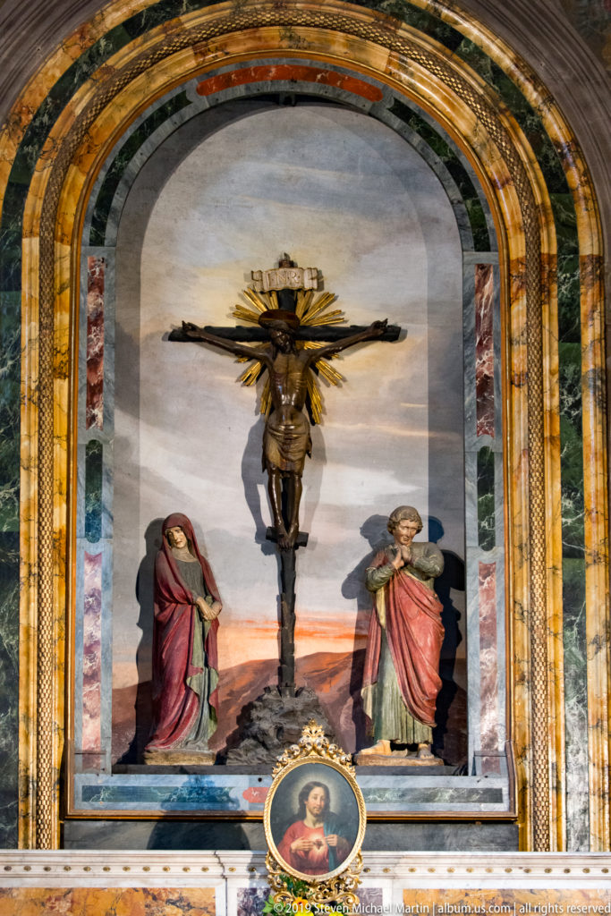 Crucifixion group with mourning figures inside an arched polychrome marble and gilded altar niche.
