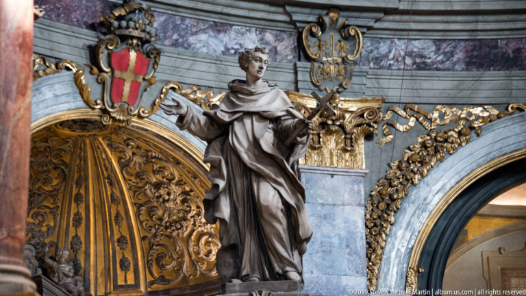Robed male saint holding a book beside gilded ornament, blue-gray stone, and a heraldic shield.