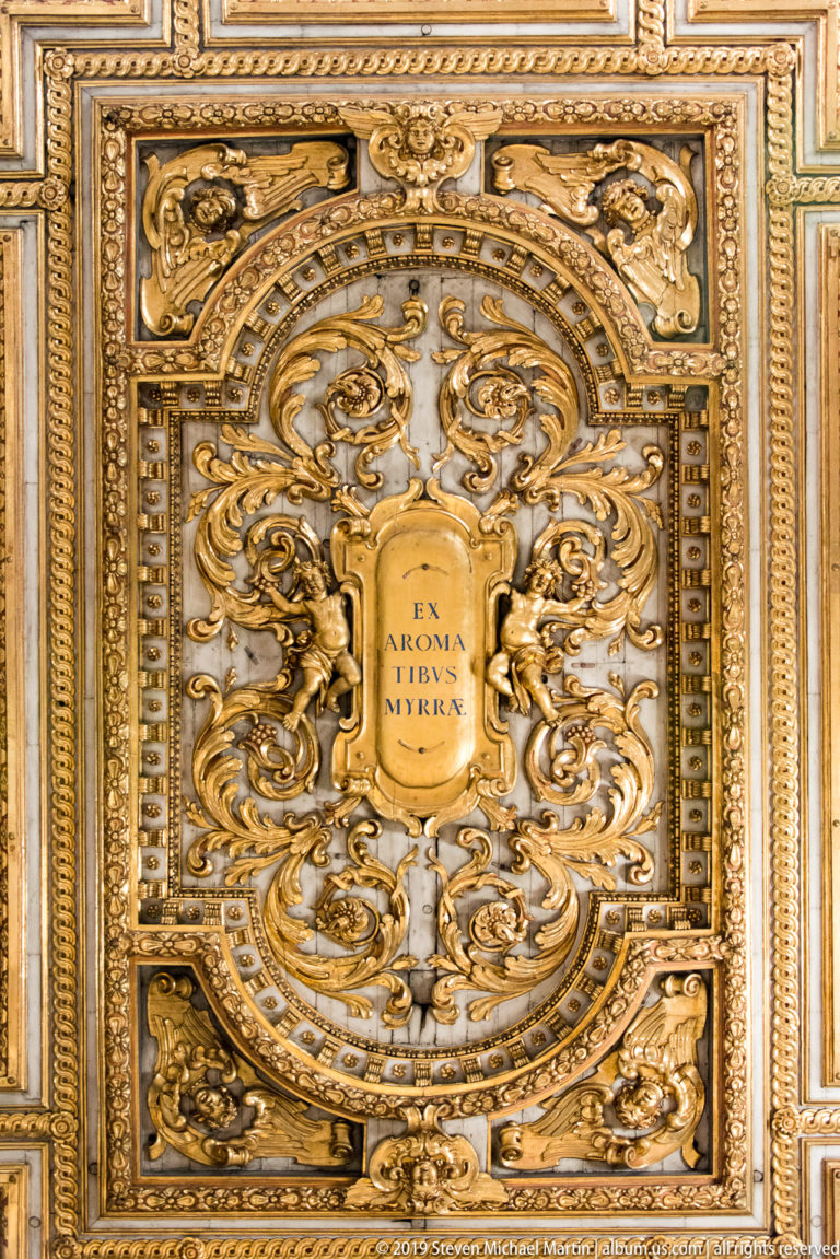 Close view of a gilded ceiling panel with scrolling relief, figures, masks, and a central Latin inscription.