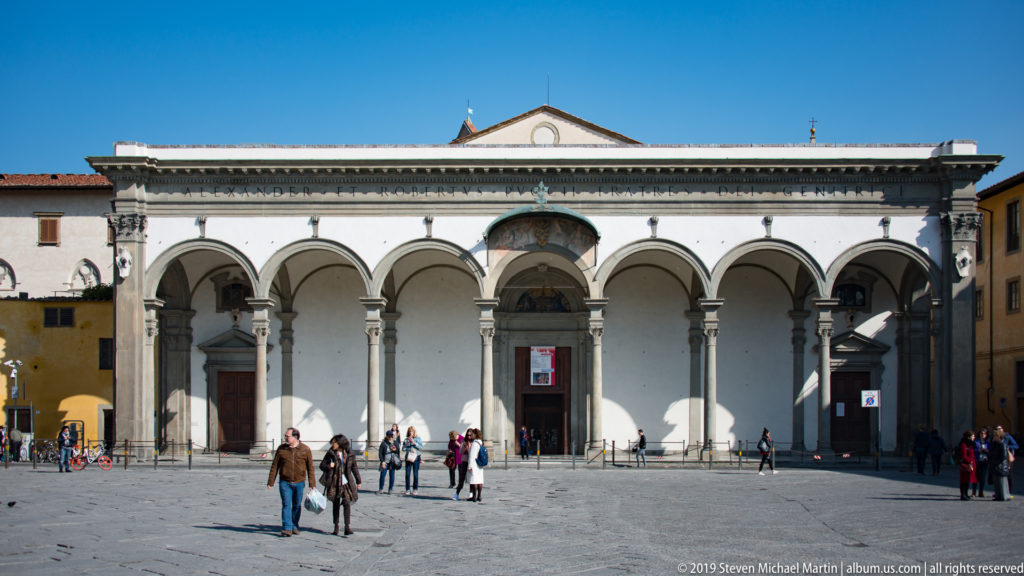 Wide exterior of Santissima Annunziata with long round-arched portico, stone columns, central pediment, and people in piazza.