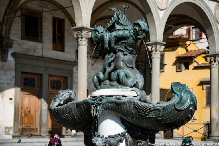 Close side view of verdigris bronze fountain with sea creatures, curving fish forms, arcade, and yellow building.