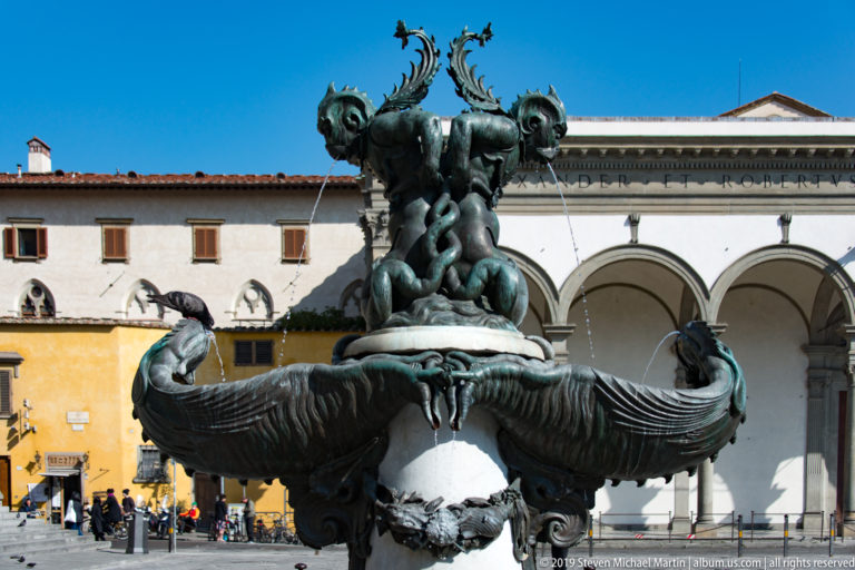 Full bronze fountain with intertwined sea creatures and sweeping fish forms before the white arcaded portico.