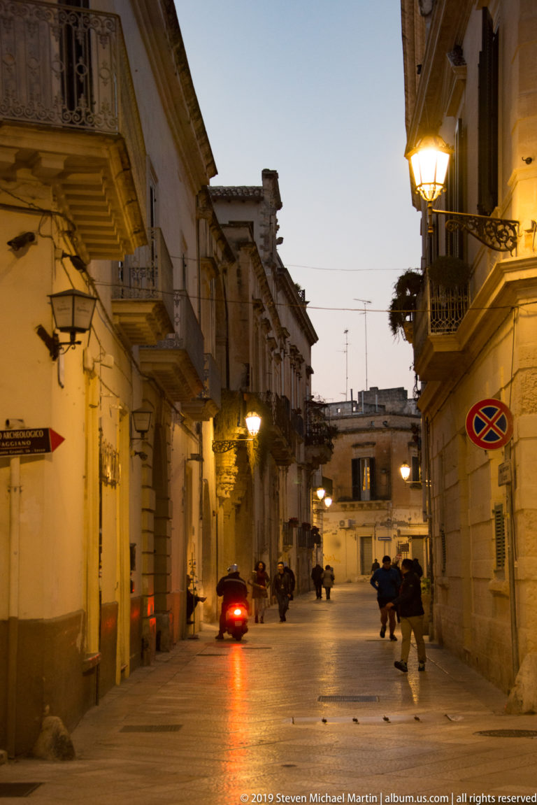 Narrow stone-paved street at dusk with balconies, glowing lamps, pedestrians, and a scooter showing a red tail light.