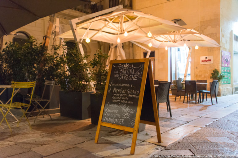 Chalkboard menu for La Cantina delle Streghe beside outdoor tables, chairs, planters, and white umbrellas on a stone-paved street.