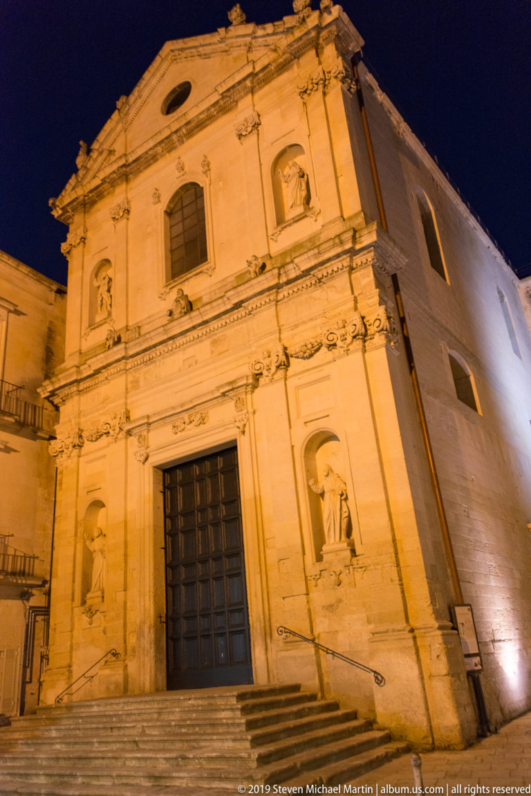Illuminated stone church façade with large wooden door, saint statues in side niches, upper windows, pilasters, and shallow pediment.