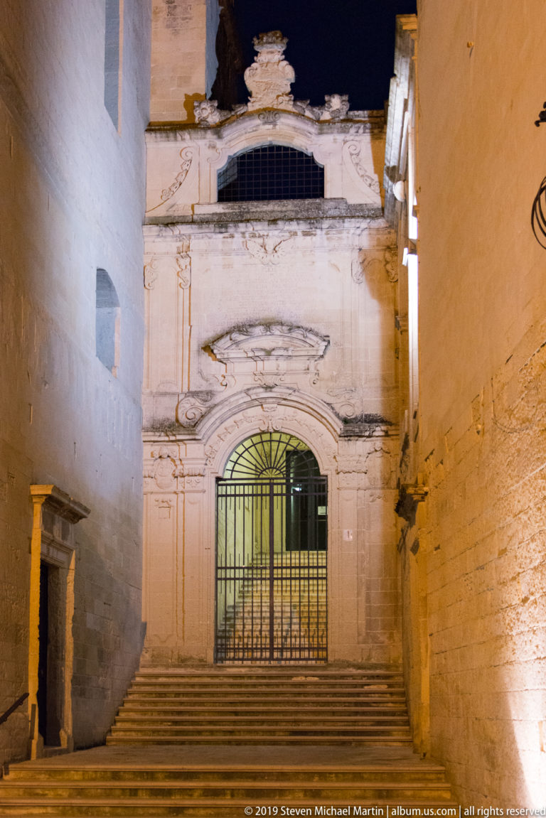 Stone steps leading to a gated arched church entrance with pilasters and a dark upper window between narrow masonry walls.