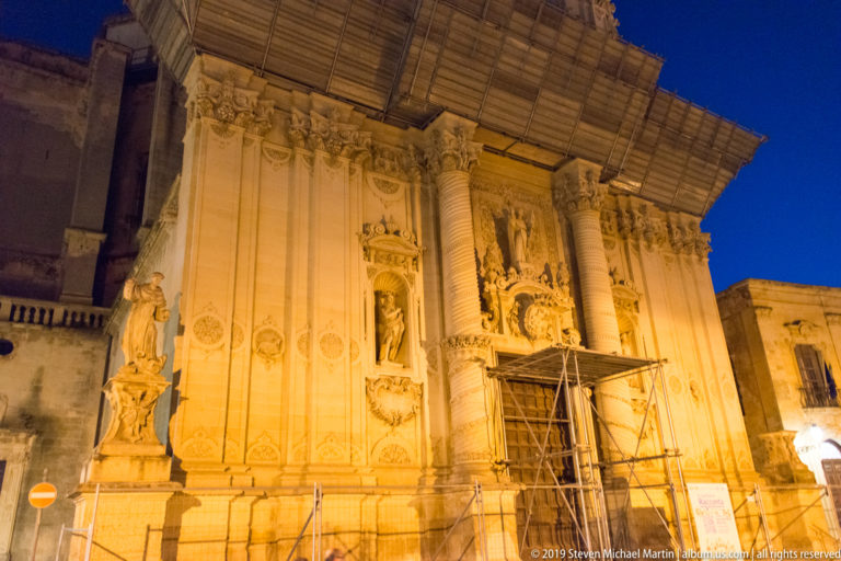Ornate illuminated church façade with spiral columns, statues in niches, carved reliefs, scaffolding, and a temporary roof.