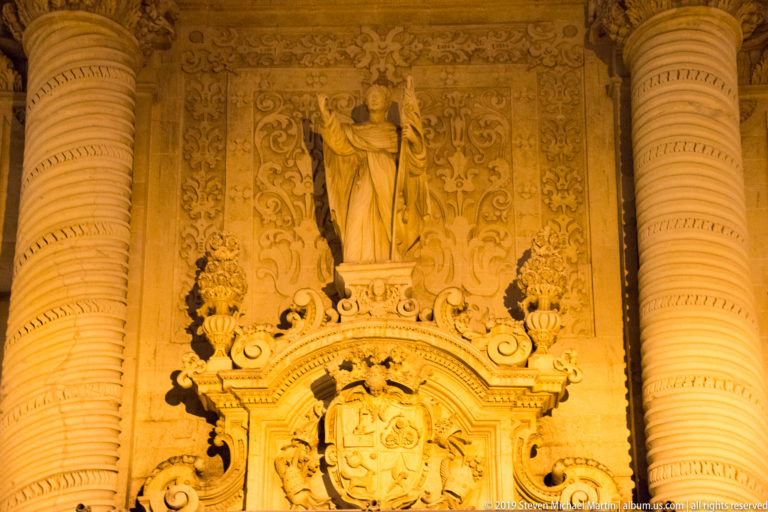 Stone statue of a saint above an ornate heraldic cartouche, framed by massive spiral columns and dense floral carving.