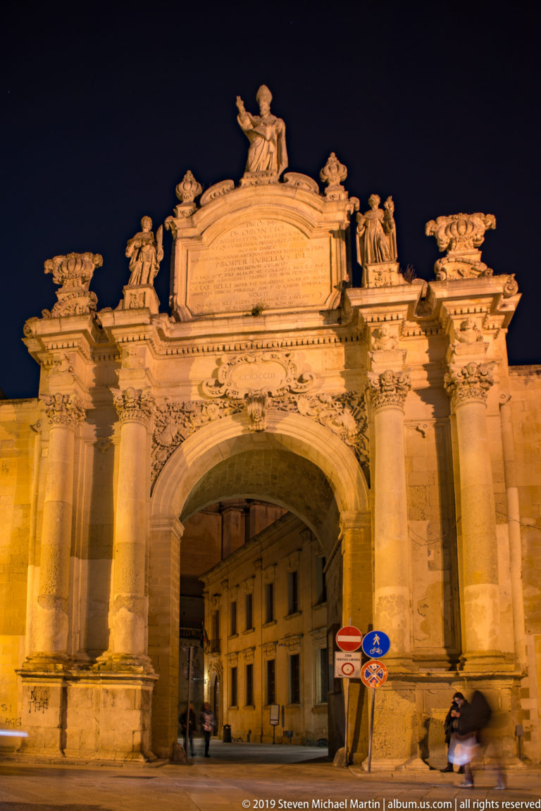 Large illuminated stone city gate with central arch, paired columns, statues, inscribed attic, and crowning figure at night.