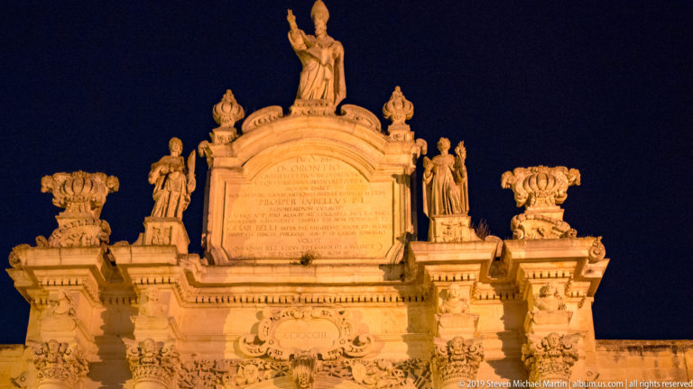 Illuminated upper section of a monumental stone gateway with inscription panel, standing statues, urns, and a central crowning figure.