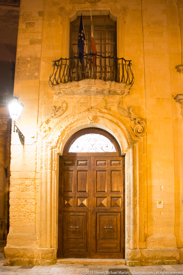 Large wooden double door in an ornate carved arch beneath an iron balcony displaying Italian and European flags.