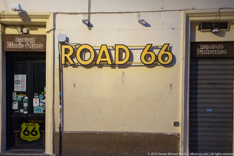 Road 66 Music & Beer storefront with large yellow-and-black sign, sticker-covered doorway, shield logo, and shuttered entrance.