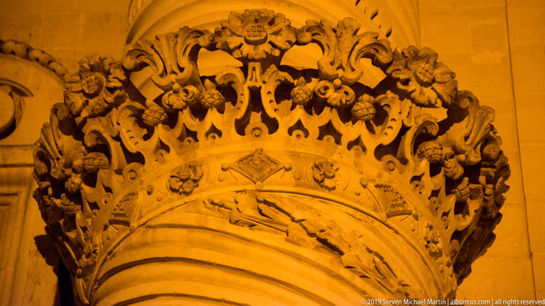 Deeply carved stone ornament above an arch with floral scrolls, masks, volutes, and crest-like motifs illuminated at night.