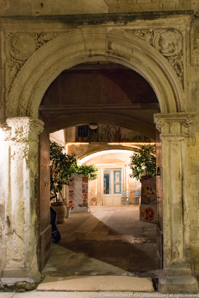 Large carved stone arch with reliefs framing a softly lit courtyard and modern doorway beyond at night.