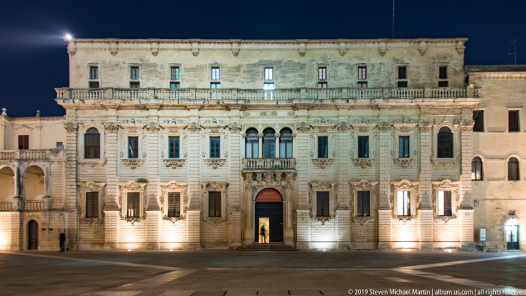 Large illuminated stone palace façade with rows of windows, carved surrounds, balustraded balcony, and central entrance in Piazza del Duomo.