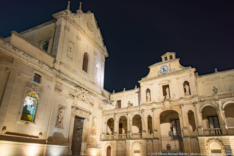 Lecce cathedral and adjoining episcopal complex in Piazza del Duomo with glowing stained glass, arcades, statues, and clock.