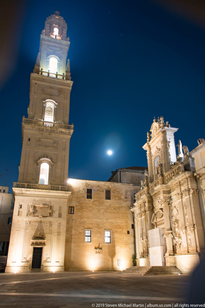 Lecce cathedral complex and illuminated bell tower enclosing Piazza del Duomo beneath a deep blue night sky and visible moon.